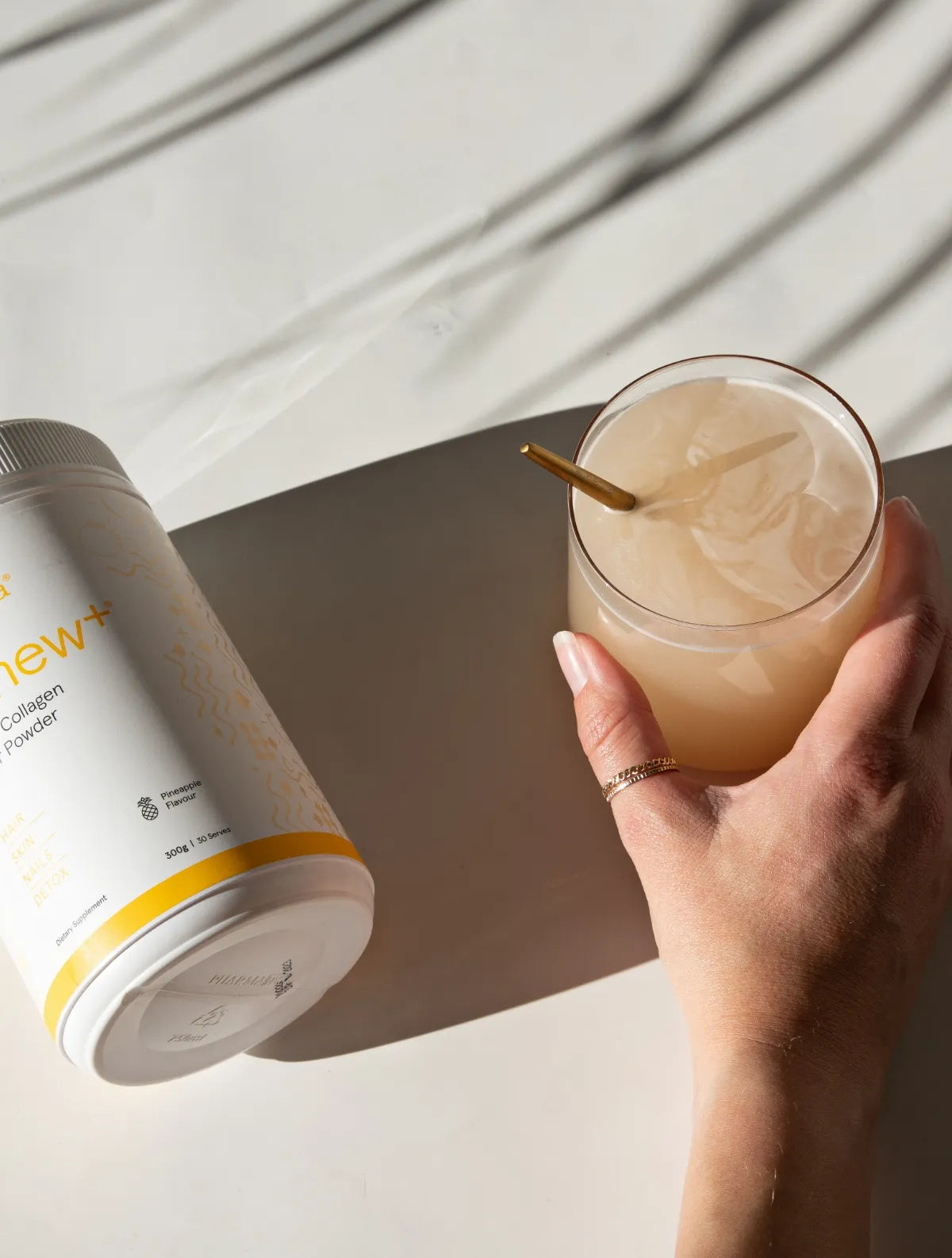 A person holds a glass of Renew+ with a gold spoon and a container of the supplement next to it.